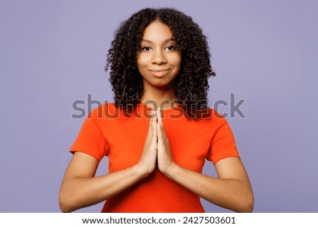 Little kid teen girl of African American ethnicity wear orange t-shirt hold hands folded in prayer gesture, begging isolated on plain pastel light purple background studio. Childhood lifestyle concept Royalty-Free Stock Photo #2427503601