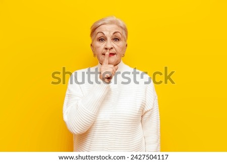 an old grandmother in white sweater holds finger near her lips asking to be silent on a yellow isolated background, an elderly woman pensioner shows a gesture of silence and secret