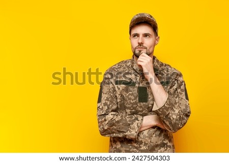pensive young Ukrainian army soldier in camouflage uniform pixel plans and thinks on a yellow isolated background, Ukrainian military cadet dreams and imagines