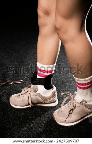 Young woman in trainers buckling a leather strap to her ankle as she sits on the floor of a gym Royalty-Free Stock Photo #2427509707