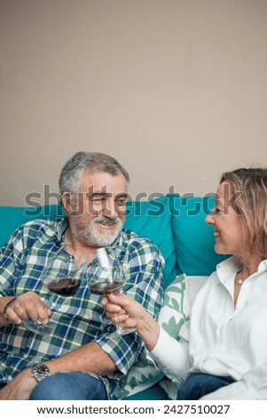 vertical portrait on their home's sofa, a senior couple shares a tender moment as they gaze into each other's eyes while raising glasses of wine in a heartfelt toast.  Royalty-Free Stock Photo #2427510027