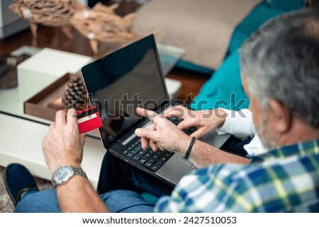 closeup from behind retired couple with red bank card in hand pointing at computer screen sitting on sofa
