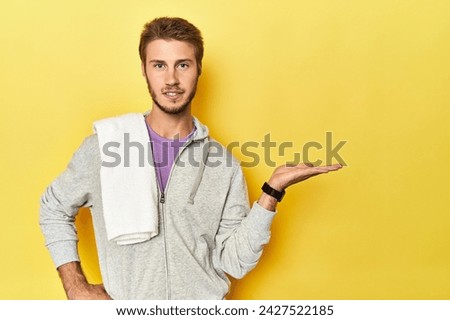 Sporty man in sweatshirt and towel on yellow showing a copy space on a palm and holding another hand on waist.