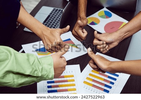 A collaborative team giving thumbs up over business performance charts on a table, indicating project approval and success