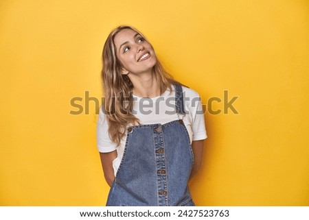 Young blonde Caucasian woman in denim overalls posing on a yellow background, relaxed and happy laughing, neck stretched showing teeth.