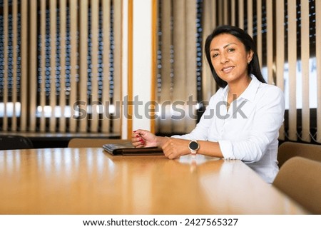 Portrait of confident positive hispanic business woman sitting with briefcase of documents at office table in meeting room.