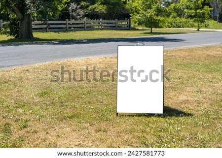 Blank white background texture of a standing board sign on the grass in a rural setting. Empty mockup template of an advertisement panel to display information or attract customers in the countryside.