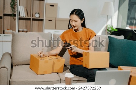 Startup small business SME, Entrepreneur owner woman using smartphone or tablet taking receive and checking online purchase shopping order to preparing pack product box.  Royalty-Free Stock Photo #2427592259