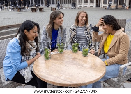 The photograph depicts a heartwarming moment among four female friends, sharing a burst of laughter at a wooden table of an outdoor cafe. Their diverse expressions of joy, from wide smiles to a hand Royalty-Free Stock Photo #2427597049