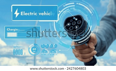 Focus EV charger pointing in front of camera display battery status hologram with blurry businessman's hand wind turbine farm. Electric car charger plug using clean energy reducing CO2 emission.Peruse