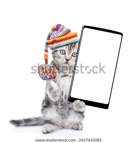 Cute kitten wearing warm knitted hat with pompon and woolen scarf holding smartphone with white blank screen in it paw. Isolated on white background