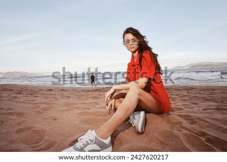 Smiling woman sitting on sandy beach, embracing the wide angle summer with trendy outfit and carefree lifestyle, radiating happiness and emotion amidst the serene sea and nature.