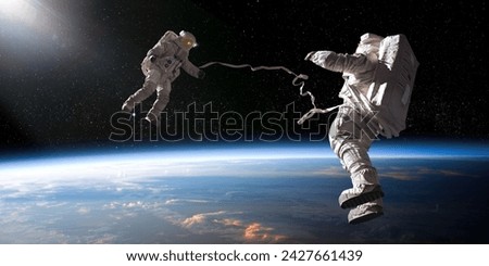 Two astronauts in full spacesuits with backpacks on a space walk with tethers, facing each other with hands out. The astronauts are in front of planet earth and some distant stars are also visible. Royalty-Free Stock Photo #2427661439
