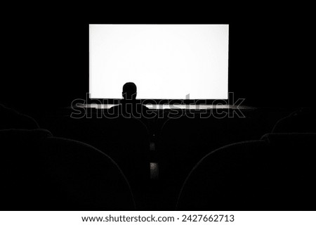 Silhouette of a man, a lone spectator sitting in a chair in the cinema hall against a white screen, rear view