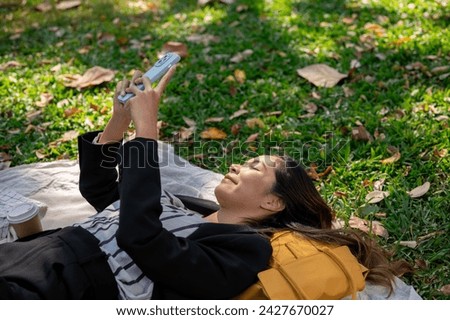 A carefree and happy Asian businesswoman is using her smartphone while lying on a picnic mat in a park, relaxing after work. people and lifestyle concepts