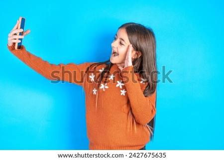beautiful kid girl wearing knitted orange sweater over blue background holds modern mobile phone and makes video call waves palm in hello gesture. People modern technology concept