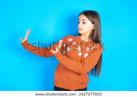 Displeased beautiful kid girl wearing knitted orange sweater over blue background keeps hands towards empty space and asks not come closer sees something unpleasant