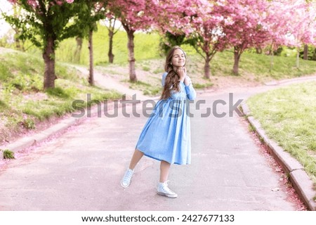 Portrait of beautiful Little girl in dress walking in blossom spring park. Young smile girl standing in blue dress at blossoming tree in the sakura garden. Beauty of woman and nature concept. 
