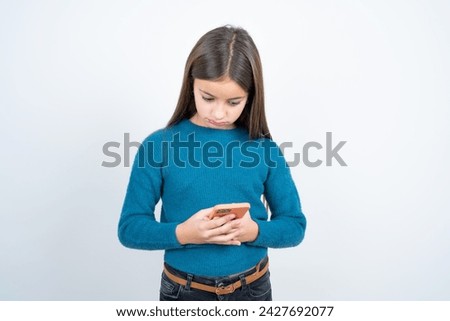 Focused beautiful kid girl wearing blue t-shirt over white background use smartphone reading social media news, or important e-mail
