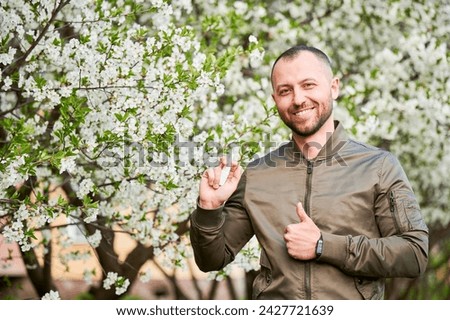 Man allergic using medical nasal drops, suffering from seasonal allergy at spring in blossoming garden. Handsome smiling man showing thumbs up near blooming tree outdoors. Spring allergy concept. Royalty-Free Stock Photo #2427721639