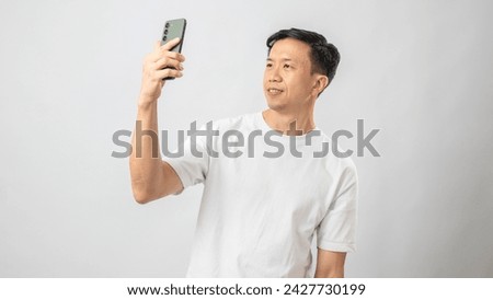 Portrait of an Indonesian Asian man, wearing a white t-shirt, taking a selfie with his smartphone and posing, isolated against a white background.