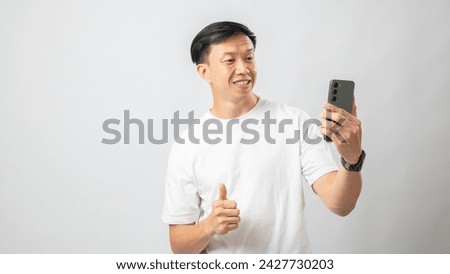 Portrait of an Indonesian Asian man, wearing a white t-shirt, taking a selfie with his smartphone and posing, isolated against a white background.