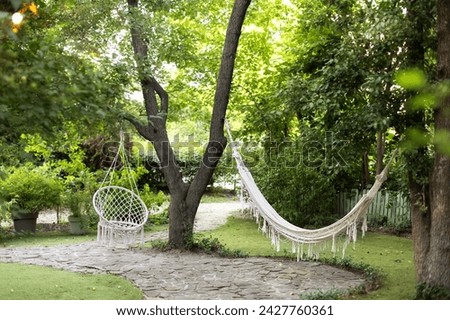 Concept of rest outdoor. Comfortable hanging wicker white chair in spring garden. Cozy hygge place for weekend relax in garden. Hammock chair in boho style hanging on tree. Cozy exterior backyard.