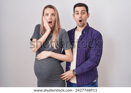 Young couple expecting a baby standing over white background afraid and shocked, surprise and amazed expression with hands on face 