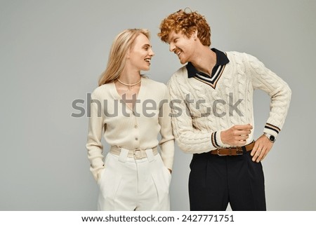 smiling and fashionable couple looking at each other on grey backdrop, timeless classic fashion Royalty-Free Stock Photo #2427771751