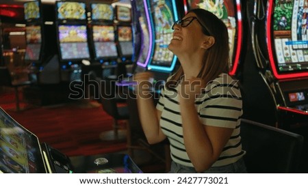 A joyful young adult woman celebrates winning at a casino slot machine. Royalty-Free Stock Photo #2427773021