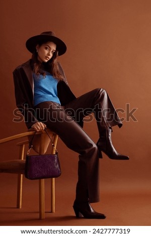 Fashionable confident woman wearing hat, blue jumper, faux leather suit blazer, pants, pointed toe ankle boots, with trendy purple bag, posing on brown background. Full-length studio fashion portrait