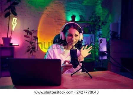 Young woman sitting in modern room with neon lights, recording video, leading online social media stream. Blogging, online services, streaming, education concept