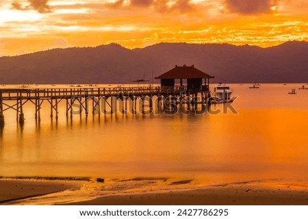 A wooden pier stretches out into a calm bay at sunset, with mountains in the background near Karanggongso Beach, Trenggalek, East Java, Indonesia.