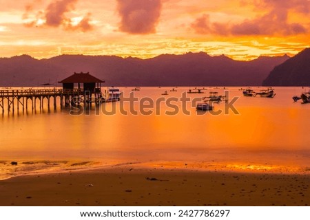 A wooden pier stretches out into a calm bay at sunset, with mountains in the background near Karanggongso Beach, Trenggalek, East Java, Indonesia.