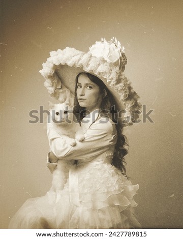 Vintage black and white conceptual photo of a girl or woman sitting with a white cat wearing a gorgeous dress and hat with a cake and burning candles on it. Birthday concept