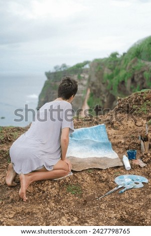 A woman artist sits on the edge of a cliff and paints a picture with acrylic paints on a canvas that lies on the ground. Landscape of the sea outdoors in summer. Drawing a picture on the ocean shore.