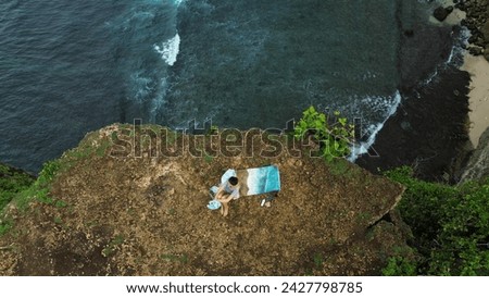 Top down view of a woman artist sitting on the edge of a cliff near the ocean and painting a picture on canvas. She paints a seascape. The process of drawing on the seashore as seen from a drone.