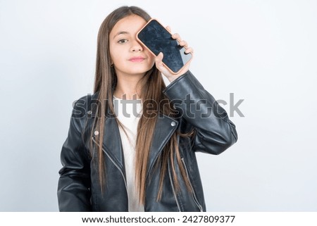 Young beautiful teen girl wearing biker jacket holding modern smartphone covering one eye while smiling