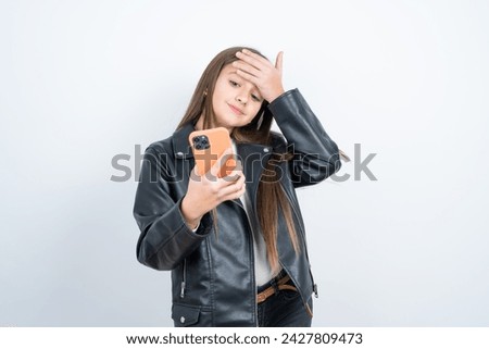 Young beautiful teen girl wearing biker jacket points thumb away and shows blank space aside, holds mobile phone for sending text messages.