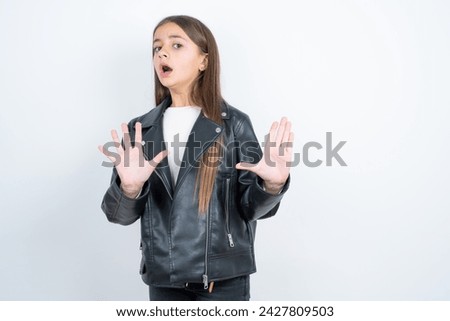 Young beautiful teen girl wearing biker jacket keeps palms forward and looks with fright above on ceiling tries to defense herself from invisible danger opens mouth.