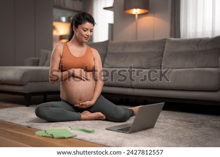 A contented young pregnant woman doing relaxing stretching exercises by following a video tutorial on her laptop in a modern, bright living room.
