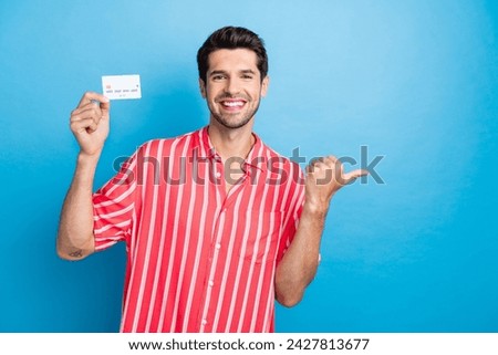 Photo of good mood cool man dressed striped shirt rising bank card pointing thumb empty space isolated blue color background