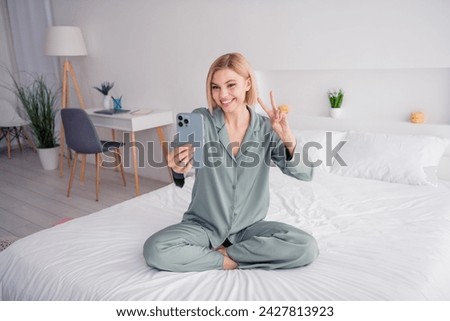 Full length photo of young blonde lady wearing gray pajama showing v sign making face portrait in camera at light white interior bedroom