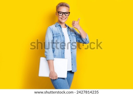 Photo portrait of pretty young girl specs hold netbook show thumb up dressed stylish denim outfit isolated on yellow color background