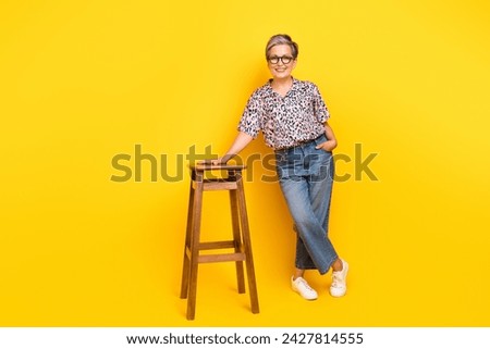 Full body photo of attractive pensioner woman stand lean on chair dressed stylish leopard print clothes isolated on yellow color background