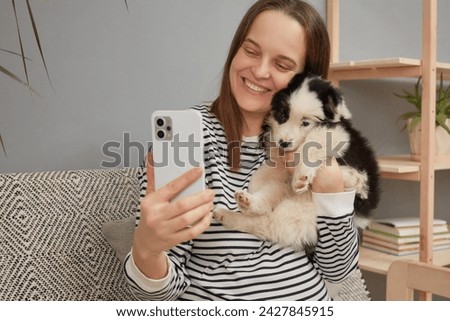Cute charming smiling young adult woman wearing striped shirt taking selfie on mobile phone with her small puppy while sitting on couch at home broadcasting livestream with her pet