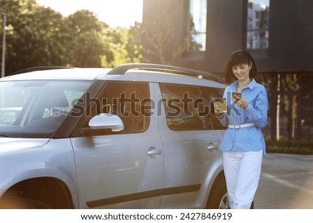 Successful businesswoman in blue shirt, white pants with paper cup of coffee, texting on smartphone while walking to the car by the modern office building. Concept of self-management for productivity.