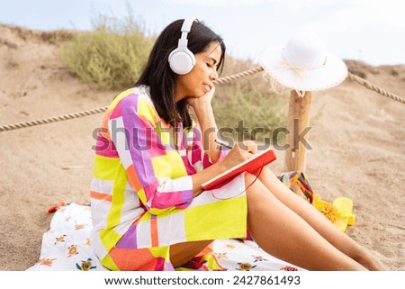 Latina woman enjoys a beautiful morning at the beach while drawing sitting on her red agenda listening to music on her headphones on a beautiful summer day
