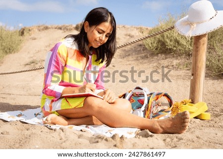Beautiful latina woman enjoys sitting on the beach relaxed while listening to music making notes and drawing in her red agenda on a beautiful summer morning