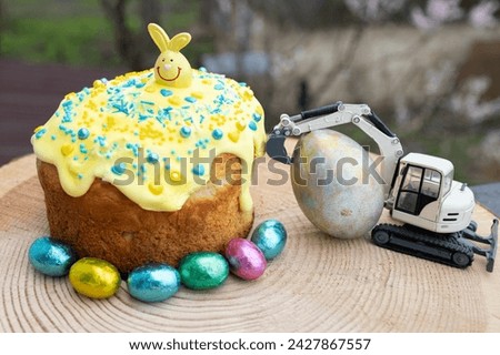 model of a toy excavator next to a large Easter egg, a holiday cake and multi-colored chocolate candies. Easter spring holiday concept, greeting card from construction business